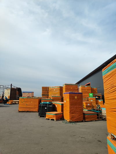 The image depicts an outdoor storage area filled with large stacks of goods wrapped in orange plastic sheeting. The materials appear to be construction supplies, possibly lumber or panels, as suggested by their uniform shapes and the pallets they are stacked on. Several tall, rectangular packages dominate the foreground and midground, while smaller stacks are visible in the background.
In the left portion of the image, a truck with an open trailer is parked. Two individuals are present near the truck. One person, wearing a blue shirt and dark pants, is standing on the ground next to a pallet, possibly arranging or inspecting the cargo. Another person, wearing a lighter colored shirt and dark pants, is standing on the trailer of the truck, looking towards the ground. A third person, wearing a blue hard hat and dark clothing, is standing on the top edge of the truck's trailer.
The scene is set against a cloudy sky, indicating either overcast weather or a transition in the day. The lighting suggests it might be late morning or afternoon. The ground is asphalt, typical of a loading dock or industrial yard. In the background, a dark gray building with a sloped roof and what appears to be an orange-framed doorway is visible. The overall impression is one of an active logistics or distribution point. No text is clearly legible on the packages or elsewhere in the image.