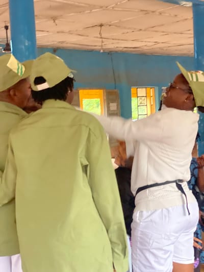 Three individuals are visible in the foreground, two wearing light green shirts and matching caps, and one wearing a white long-sleeved shirt and white shorts. The person in white is facing away from the viewer, with their right arm extended towards the person on the left in the green shirt. The person on the left in the green shirt is also facing away from the viewer, their left shoulder visible. The third individual, also in a green shirt and cap, is partially obscured between the other two. The environment appears to be an indoor space with a light-colored, textured ceiling and blue walls. Two windows with orange frames are visible in the background, through which greenery can be seen. The scene takes place in Ise-Ekiti, Nigeria.