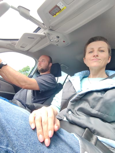 In the foreground, a person's hands, with polished pink fingernails, rest on their lap covered in blue jeans. Next to them, a woman with short brown hair and a light blue top underneath a grey and light blue puffer jacket is visible, looking towards the camera with a slight smile. Her seatbelt is fastened. In the background, a man with a beard and a dark t-shirt is driving, also wearing a seatbelt. The scene takes place inside a car with a sunroof open. The car appears to be on a road, with trees visible through the window. The lighting suggests it is daytime. The car is likely in the vicinity of Фаніпальскі сельскі Савет, Belarus, given the user's prompt specifying this location.
