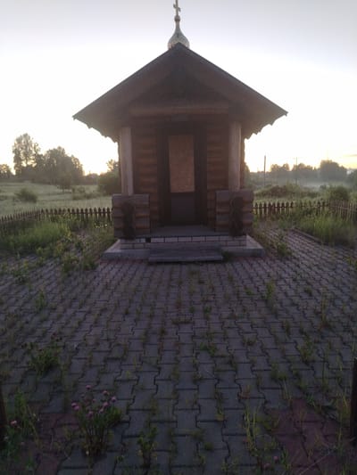 A small, wooden chapel or shrine stands centrally in the image, facing the viewer. The structure is built from horizontal logs, stained or aged dark brown, and features a steep, dark-colored pitched roof. At the apex of the roof is a golden, onion-shaped dome surmounted by an Orthodox cross. The front entrance of the chapel is covered by a light-colored wooden panel or board. The structure rests on a base of masonry blocks, topped by a stone slab that forms a small platform or step. Two decorative, log-like columns flank the base of the entrance.

The foreground is paved with interlocking dark gray concrete pavers, through which numerous green grasses and small plants, including some with small pink-purple flowers, are growing. This indicates a lack of recent maintenance.

Behind the paved area, a low, rustic wooden picket fence runs horizontally across the mid-ground, showing signs of weathering and some disrepair. Beyond the fence is an unkempt grassy field or meadow with scattered trees. The background features a line of denser trees, largely silhouetted against a bright, hazy sky. The sky appears overcast or illuminated by a low sun, possibly indicating dawn or dusk. A faint mist or haze is visible in the distant background, particularly over the field. On the far right horizon, faint structures resembling utility poles or power lines are discernible. No individuals or animals are present in the scene. The location depicted is Petrovskoye, Russia.