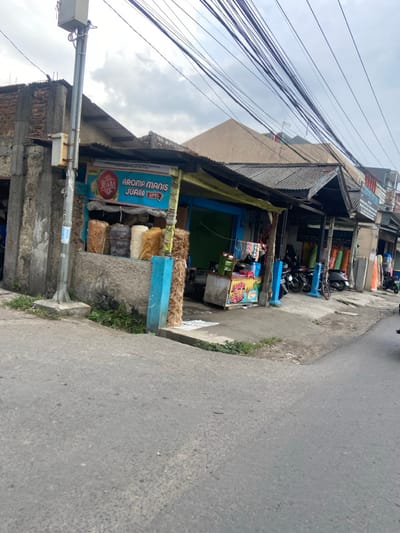 The image shows a street corner in Kecamatan Cimahi Selatan, Indonesia. A small shop with a blue awning and a sign that reads "Aroma Manis Juara Apel" is located on the corner. The shop is selling a variety of goods, including rolls of fabric and other merchandise. There are several motorcycles parked in front of the shop. The street is paved and there are power lines overhead. There are no people visible in the image. The image was captured during a cloudy day. The image was taken at the coordinates -6.93 latitude, 107.565 longitude.