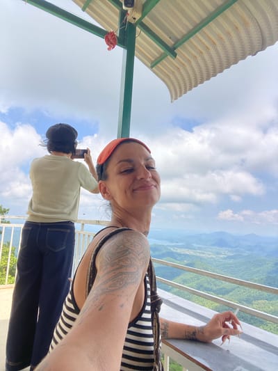 The image captures two individuals on a balcony overlooking a vast, mountainous landscape, likely in or near Phan Son, Vietnam, given the context of the question. The foreground features a woman, looking towards the camera with a smile and her eyes partially closed. She is wearing an orange headscarf and a black and white striped tank top. Her right arm is resting on a table, and she holds a cigarette between her fingers. She has visible tattoos on her arm and shoulder. To her left, a second person stands with their back to the camera, wearing a light green long-sleeved shirt, dark blue jeans, and a dark beret. This person is holding a smartphone, appearing to take a photo of the scenery. The balcony has a metal railing and a concrete table. Above them, a corrugated metal roof is supported by green beams. The background reveals a picturesque view of rolling green hills, a body of water, and distant mountains under a cloudy sky.
