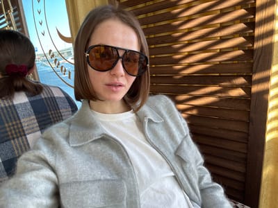 The image depicts an indoor scene in Mysiadło, Poland, featuring a female subject facing the camera in a medium shot. She has short brown hair and wears large aviator-style sunglasses with orange-tinted lenses, a white undershirt, and a light grey textured, zippered jacket with a collar and two chest pockets. Her lips are slightly parted. To her left, the back of a second individual's head is partially visible, showing brown hair secured with a dark red scrunchie and a blue, white, and brown plaid garment. The background on the left reveals an outdoor parking area through a window, displaying patches of snow, several vehicles, and a blue "P" parking sign. A large, ornate, gold-colored decorative element is visible through the window. The background on the right is a wooden slatted panel with horizontal sunlit shadows, and a narrow strip of yellow fabric, possibly a curtain, on the far right edge.