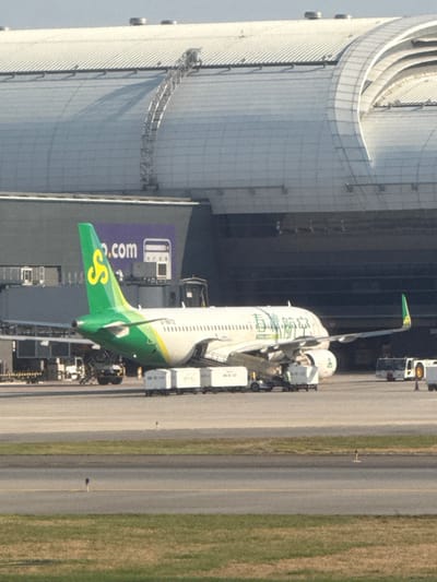 An Airbus A320-200 aircraft, bearing the registration B-6972 and painted in Spring Airlines livery (white fuselage with a green tail featuring a yellow 'S' logo, and Chinese characters on the forward fuselage), is parked on an asphalt airport apron under clear daylight conditions. The aircraft is positioned with its tail section towards the left of the frame, angled slightly away from the viewer.
To the left of the aircraft, a line of six white baggage or cargo container carts are aligned near the aircraft's fuselage, with a small ground service vehicle partially visible behind them. Another larger, yellow-and-white ground support vehicle, possibly a pushback tug, is positioned to the right of the aircraft, behind its right wing. No individual personnel are distinctly visible, but ground operations are implied by the presence of the equipment.
In the background, a large, modern airport terminal building is visible. It features a distinctive curved, light-colored metallic roof structure with multiple arched ribs and a dark grey lower section. A prominent purple banner or sign on the dark section displays "o.com" and a white graphic resembling a fuel pump or payment terminal, consistent with "trip.com". Elevated walkways or maintenance structures are visible on the roof.
The immediate foreground consists of a strip of dark green grass with small blue markers, transitioning to a grey asphalt surface, indicating a taxiway or runway beyond the apron where the aircraft is parked. The scene is located within Racha Thewa Subdistrict, Thailand, consistent with Suvarnabhumi Airport.