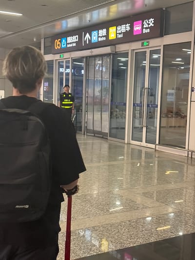 A person with blonde hair, wearing a black shirt and backpack, is seen from behind, holding a red suitcase handle. They are in an indoor environment, likely an airport or train station, with a polished, reflective floor. In the background, a sign displays "05 Arrival Exit," "Metro," "Taxi," and "Bus" in both Chinese and English, indicating transportation options. A security guard wearing a high-visibility vest is standing near the entrance. The location is identified as Xinwu District, China.
