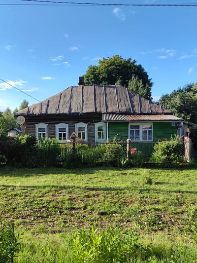 The image shows a rustic, weathered wooden house with a dark, tarp-covered roof. The house has several windows with white trim and curtains visible. The front of the house is partially covered by dense green foliage and a low fence. To the left of the house, a satellite dish is mounted on a wooden pole. To the right, a section of the house is painted green, and a house number "21" is visible. The scene appears to be in a rural or suburban setting, possibly a dacha or village home, with lush green grass in the foreground and trees in the background. The sky is blue with a few wispy clouds. The location is identified as Квант-88 Снт, Russia. There are no people or animals visible in the image, and no actions or interactions are depicted. The overall impression is of a quiet, somewhat aged, residential property.
