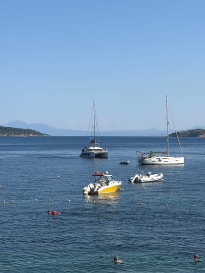 A wide-angle, eye-level shot captures a serene coastal scene in Skiathos, Greece. The dominant feature is the deep blue, rippling water of the Aegean Sea. Several boats are anchored: a white catamaran with a tall mast is prominent, accompanied by a smaller white inflatable boat. Further in the distance, another boat with a yellow section is visible. A person in an orange life vest and shorts is swimming near a line of yellow buoys. The background features a hazy, mountainous coastline covered in dark green vegetation, with a clear, bright blue sky above. The time of day appears to be midday, with bright sunshine casting a gentle sheen on the water and indicating fair weather. The overall mood is tranquil and relaxed, characteristic of a summer day at a Greek island.