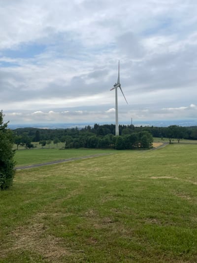 A solitary wind turbine stands tall against a cloudy sky. The turbine, with its white tower and three blades, is positioned on a gentle slope overlooking a verdant landscape. To the right of the turbine, a green field stretches out, bordered by a thin, winding path. Beyond the field, a line of trees creates a dark, dense band against the horizon. The sky is a mosaic of white and grey clouds, with patches of pale blue peeking through. The overall impression is one of a peaceful, rural setting. The image appears to have been taken during the daytime, under overcast or partially cloudy weather. The scene suggests a remote, possibly agricultural area, with the wind turbine indicating a focus on renewable energy. No text is visible in the image. The orientation of the image is portrait.