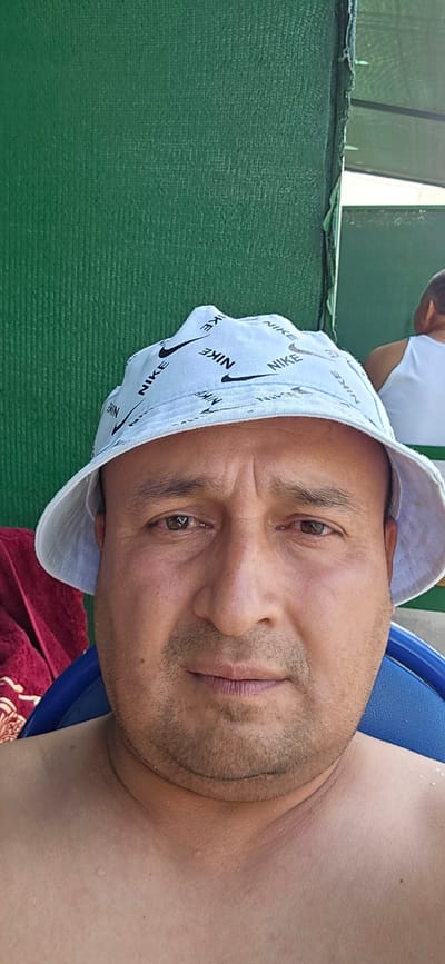 A man wearing a light blue bucket hat with a repeating Nike logo and swoosh pattern is shown from the chest up. The hat is tilted forward, obscuring his forehead. He has short, dark stubble on his face and appears to have brown eyes. He is looking towards the camera. The background is a textured green surface, possibly a mat or fabric. To the left of the man, there is a glimpse of a blue chair and a reddish-brown textured cloth with a pattern. The man's shoulders are bare and appear slightly pink, suggesting sun exposure. There are small water droplets visible on his skin. The lighting suggests it is daytime. The overall impression is of a casual outdoor setting.