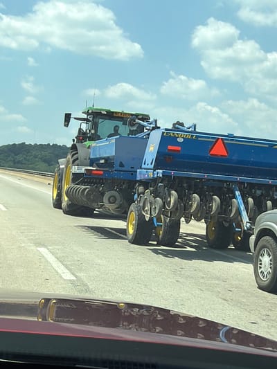 A green and black tractor, possibly a John Deere, pulls a large blue agricultural implement with the brand name "LANDOLL" visible in yellow lettering. The implement is equipped with multiple seeding or planting units, each with small wheels. The tractor and implement are traveling on a paved highway, indicated by lane markings. In the background, a dense line of trees forms the horizon, against a sky with scattered white clouds. The scene appears to be in a rural or semi-rural area, given the agricultural machinery and natural landscape.