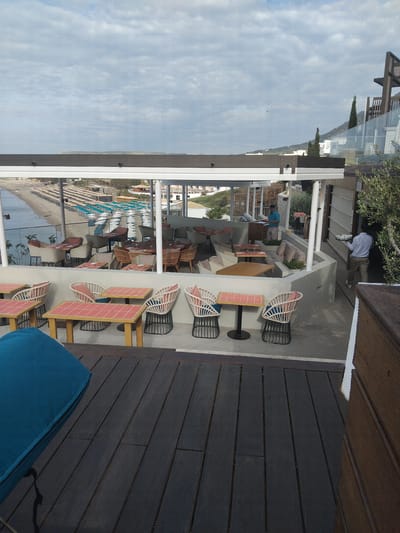 Captured from an elevated vantage point, the image showcases an outdoor restaurant or bar space in Pyli, Greece. The restaurant area has tables with coral-colored tops and white chairs. It is partially enclosed by a structure with reflective glass panels. Outside the enclosure, the landscape includes a beach with lounge chairs, the sea, and the sky. A man stands under a pergola and is carrying dishes.
