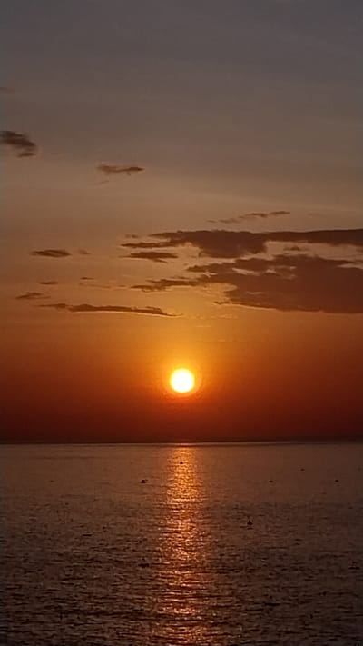 The image depicts a sunset over the ocean. The sun, a bright orange orb, is low on the horizon, casting a warm glow across the sky and the water. The sky is a gradient of colors, from a pale blue at the top to a deep orange near the horizon, with scattered clouds. The ocean is calm, with gentle ripples reflecting the colors of the sunset. The location is the coastline of Tuy Phong District, Binh Thuan Province, Vietnam, known for its beautiful beaches and stunning sunsets. There are no people or specific objects visible in the immediate scene, just the natural elements of the sky, sun, and sea.