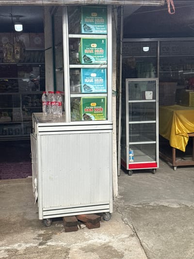 The image shows a street-level view of a small shop in Lampisang, Indonesia. The shop appears to be selling food products, specifically a type of cake called "Kue Adee Meureudu" by a brand named "KAK NUR". Four boxes of this product are prominently displayed in a glass-fronted display case. The boxes feature images of the cake and text in Indonesian, including "Selamat Menikmati" (Enjoy) and "Terima Kasih" (Thank you).

Behind the food display, there's another glass display case with a red trim, which appears empty except for what looks like a can or bottle inside. Further back, a yellow tarp is draped over something, suggesting storage or a temporary structure. The shop itself is constructed with metal framing and glass, with a roller door visible on the right side of the image.

The ground is concrete and somewhat worn, with a few bricks supporting the display unit. A set of plastic water bottles is also visible in front of the display case. The lighting suggests it is daytime, possibly late morning or early afternoon, with bright sunlight casting shadows. The overall impression is of a modest, functional storefront in a local Indonesian community. No people are directly visible in the frame, but the setting implies ongoing commercial activity.