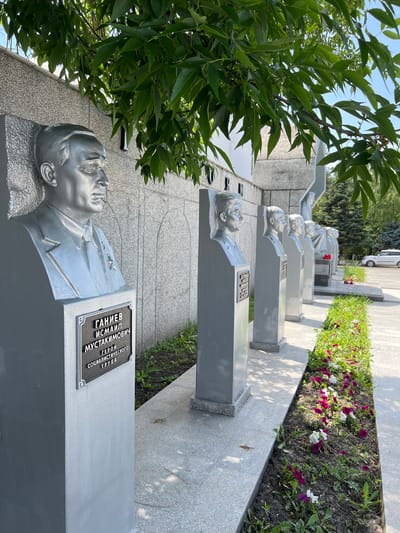 In the city of Takhtamukai, Russia, a row of silver busts of men is displayed outdoors under the shade of lush green trees. The busts are mounted on rectangular grey pedestals, each bearing a dark plaque with engraved text. The first bust, closest to the viewer, is of a man with a stern expression, wearing a suit and tie, and adorned with medals. Its plaque reads "ГАНИЕВ ИСМАИЛ МУСТАКИМОВИЧ ГЕРОЙ СОЦИАЛИСТИЧЕСКОГО ТРУДА" (Ganiew Ismail Mustakimovich, Hero of Socialist Labor). Behind this bust, a series of other busts are visible in a receding line, though their details are less clear. The busts are arranged along a paved walkway, bordered by a flower bed filled with blooming pink and white flowers. In the background, a textured grey wall and trees suggest a public space or monument. A white car is parked further down the path, and the sky is a clear blue.