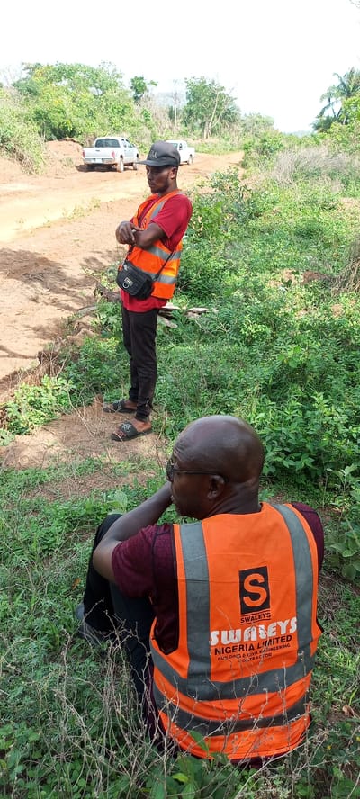 Two men are depicted outdoors in what appears to be a rural or undeveloped area in Otunade, Nigeria. The scene features lush green vegetation, dirt tracks, and scattered trees under a bright, hazy sky.

In the foreground, a bald man with glasses is sitting or crouching, viewed from behind and the side. He wears a maroon t-shirt and an orange reflective vest. Visible on the back of his vest is a black logo featuring a large 'S', with the text "SWALEYS" above "SWALEYS NIGERIA LIMITED BUILDING & CIVIL ENGINEERING CONTRACTOR". He is looking towards the right side of the frame, seemingly resting or observing.

Behind him, another man stands upright, his arms crossed. He wears a red t-shirt, an identical orange reflective vest, black pants, and dark sandals. A black cross-body bag hangs across his chest. He also wears a black cap with the letters "OLO" visible on it. His gaze is directed to his right, giving an impression of observation or contemplation.

In the background, on a dirt road, two pickup trucks are parked or driving slowly. One appears silver/grey, and the other is white. The presence of the reflective vests and the company name on them suggests that the men are likely involved in surveying, construction, or civil engineering work in this undeveloped locale. The abundant greenery and dirt pathways indicate an environment that is not highly urbanized. The time of day appears to be bright daylight.
