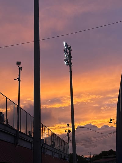 This image captures an outdoor stadium or sports complex during sunset in San Antonio, Costa Rica. The sky displays a gradient of colors, transitioning from vibrant orange and gold near the horizon to shades of purplish-pink overhead, with scattered clouds illuminated by the setting sun.

In the foreground and midground, multiple tall, dark poles are visible. A prominent, tall, slender pole is positioned slightly left of center, extending out of the frame at the top. To its right stands another equally tall, slender pole, topped with a cluster of five illuminated stadium floodlights emitting bright white light. Further to the left, a shorter pole supports a single floodlight at its apex and several smaller, dark camera-like or sensor devices mounted below it. On the lower right, a partial view of a fourth, shorter pole with two speaker-like attachments is visible.

A dark, chain-link fence, likely part of a spectator area or bleachers, extends diagonally across the left side of the frame. Below the fence, a section of a structure with a reddish-brown roof is discernible. Silhouetted figures, approximately five to seven individuals, are visible behind this fence, appearing to be seated or standing on the spectator structure. Their features are indistinguishable due to backlighting.

A thin, dark horizontal wire traverses the upper portion of the sky. In the lower right quadrant, beyond the main structures, silhouetted buildings and multiple crisscrossing power lines are visible against the lighter sky, suggesting an urban or semi-urban setting. The overall scene is characterized by the strong contrast between the illuminated sky and the dark, silhouetted foreground elements.