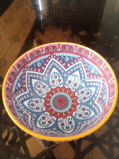 A decorative, brightly colored ceramic bowl with a yellow rim sits on a dark, reflective surface. The bowl features a intricate mandala-like pattern in shades of blue, red, and white. The surface appears to be a table with a geometric pattern visible through its reflection. There are no people or active events depicted in the image. The scene is static and focuses solely on the bowl and its immediate surroundings. The location is not specified in the image itself.