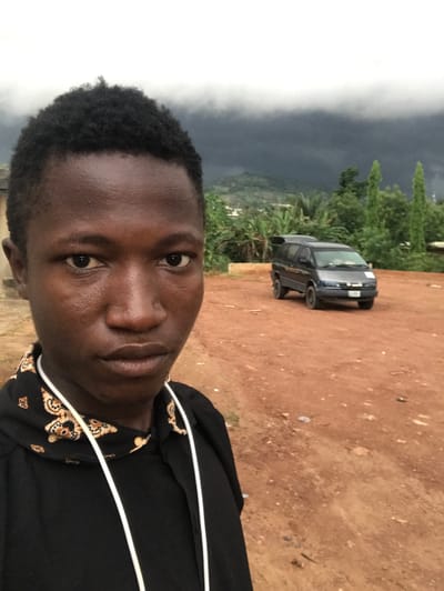 A close-up self-portrait of a young, dark-skinned man, taken outdoors. He has short, dark curly hair. The background shows a dark-colored sedan parked on a reddish-brown dirt surface. Further in the background, there are green trees and a cloud-obscured horizon, likely indicating a mountainous or elevated area. The man is wearing a dark garment with a patterned collar and white drawstrings. The location is potentially in Ife, Nigeria, though this cannot be definitively confirmed from the image alone.