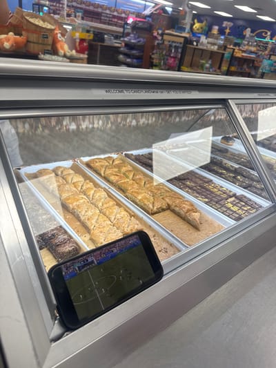 The image captures a display case filled with various sweets, likely in a confectionery or dessert shop located in Ocean City, United States of America. The display case features rows of baklava, chocolate bars with gold leaf accents, and other confectioneries. In the foreground, a smartphone is propped up, displaying a soccer match. The background reveals a store environment with shelves stocked with merchandise, possibly candy or novelty items, and some character figures. The overall lighting suggests an indoor setting, likely a brightly lit commercial establishment.