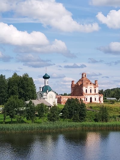 This is a vertical, eye-level, medium-shot photograph of a rural landscape on a sunny day with a blue sky and white clouds. In the foreground, a body of dark blue water with ripples reflects the sky and a bit of the bank. The bank is lined with tall green reeds. Behind the reeds, a grassy slope leads up to several structures.
On the left side of the slope, a white building with a green dome and a cross on top is partially visible, surrounded by trees. This is likely a church. To the right of this building, a red-brick ruin of another building, also appearing to be a church, stands prominently. It has a collapsed roof and exposed brickwork, suggesting it is abandoned. The ruined building has arched windows and decorative elements.
Further back, a line of green trees forms the horizon, with a few smaller structures and fields visible beyond them. The overall scene depicts a peaceful, rural setting with historical architecture. The presence of two churches, one in good condition and one in ruins, suggests a historical or religious site. This is likely the village of Белоусово, Russia, which is known for its churches.