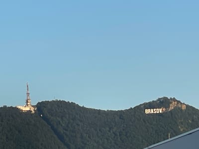 The image shows a wide shot of a forested mountain range under a clear blue sky. On the left side of the mountain, a tall, multi-tiered radio tower stands on a building complex. Further to the right, on another peak, the word "BRASOV" is illuminated in large, block letters. The base of the mountain range appears to be covered in dense green trees. In the foreground, a portion of a dark, angled rooftop is visible, partially obscuring the view of the mountain. The scene appears to be a landscape view from the city of Cristian, Romania, likely taken during daylight hours.