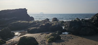 The image shows a rocky shoreline with the sea in the background. The foreground consists of large, dark rocks, some covered in moss, interspersed with sand and small pebbles. The sea is a deep blue with gentle waves. In the distance, on the horizon, a small, conical island can be seen. The sky is hazy and light, suggesting it may be early morning or late afternoon. The overall scene depicts a natural coastal landscape. This is likely the Ailsa Craig island, visible from Girvan, United Kingdom.