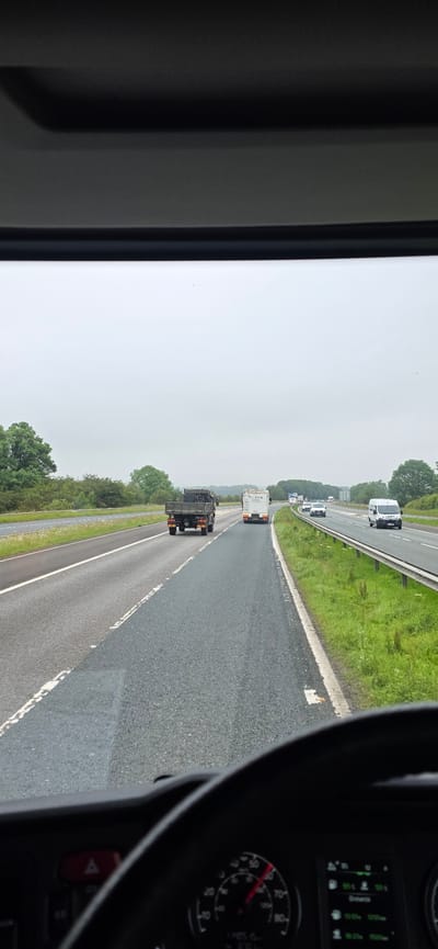 The image is a first-person view from the driver's seat of a vehicle, looking out through the windshield at a highway. The sky is overcast and the weather appears to be dull. The road is asphalt with white lane markings. In the foreground, the dashboard and steering wheel of the vehicle are visible. The steering wheel is black and a speedometer is partially visible, showing a speed of approximately 60 mph.

On the highway, there are several vehicles ahead. The closest vehicle is a dark-colored truck with an open bed. To the right of this truck is a white box truck. Further ahead on the right lane, there is a white van. On the left side of the highway, there are trees and green grass. The environment suggests a rural or suburban setting on a motorway. There are no visible individuals. The specific location in the city of Barton, United Kingdom, cannot be determined from the visual information provided.