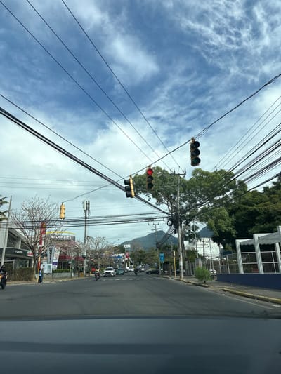 The image shows a street intersection in San Rafael, Costa Rica (latitude: 9.937, longitude: -84.152). The scene is captured from a car traveling through the intersection. There are numerous power lines crisscrossing the sky, obscuring the view of a blue sky with white clouds. There are two sets of traffic lights: one with a red light and another with a green light. Several vehicles, including cars and motorcycles, are in motion.  A sign for McDonald's is visible on the left side of the road, and there are trees lining the streets. The environment appears to be a typical urban area with a mix of residential and commercial buildings.