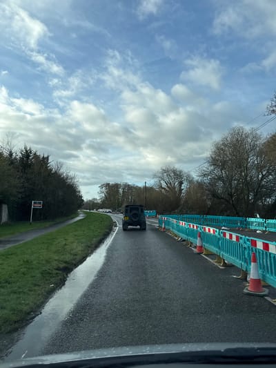 The image displays a rural road scene in New Forest, United Kingdom, observed from the interior of a vehicle looking forward on an overcast day. The asphalt road surface is wet and reflective, indicating recent precipitation, with a substantial puddle running along the left edge of the carriageway. A dark-colored Land Rover Defender, equipped with a rear-mounted spare wheel, is positioned centrally in the frame, traveling away from the viewer.
To the right of the road, a construction or maintenance area is demarcated by a continuous line of interlocking turquoise plastic barriers with red and white reflective panels. Several orange and white traffic cones are positioned immediately in front of these barriers. The ground beyond the barriers appears disturbed. Bare deciduous trees and overhead utility wires are visible on the right side further in the background.
On the left side of the road, a green grass verge borders a paved pedestrian pathway. A red "FOR SALE" sign with white text is affixed to a white pole on the grass verge. Dense evergreen and deciduous trees and shrubs line the property boundary behind the pathway. In the distant background, a line of light-colored vehicles is discernible along the road as it curves gently to the left. The sky is partially covered with white and grey clouds, revealing sporadic patches of blue. No individuals are visible in the scene.