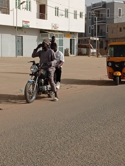 In the city of Gayawa, Nigeria, a daylight street scene shows two males on a dark-colored motorcycle positioned on a paved road. The driver, wearing a grey agbada-style garment and a patterned cap, has his right hand raised to his ear, smiling. He wears light-colored sandals. Seated directly behind him is a male passenger in a white t-shirt and dark pants. A third male, wearing a red shirt, is visible standing behind the motorcycle. To the right, an orange three-wheeled auto-rickshaw (Keke Napep) with "TFA 05" partially legible on its front is present. The road surface is granular and appears slightly dusty. In the background, a multi-story white building with green-framed windows and a visible sign reading "SUKIYA SHOP" stands to the left. Further right in the background, another multi-story building with a grey facade and white trim is visible. The scene is brightly lit by natural sunlight, casting sharp shadows.