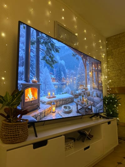 An interior shot captures a cozy living space, likely in the evening, with a large flat-screen television as the focal point. The TV displays a serene, animated winter scene featuring a snowy landscape with cabins and a tranquil body of water under a twilight sky, viewed from a stone patio. On the patio, a lit fireplace, hurricane lamps with candles, and a pet bed with a sleeping animal (possibly a cat or small dog) create a warm ambiance. A table on the patio holds drinks and snacks.
In the physical room, warm fairy lights are strung across the plain light-colored wall, adding to the inviting atmosphere. An adjacent wall features textured stone-like wallpaper. A simple rectangular, silver-framed piece of art hangs on the wall beside the TV. The television rests on a white, modern TV stand or cabinet, which has open compartments. Inside one compartment, a decorative bookstand holds an open book, and another compartment contains a woven decorative box. A green houseplant in a woven basket sits on the floor to the right of the TV, and another leafy plant is visible behind the TV on the cabinet. No people are visible. The overall mood conveyed is one of relaxation and comfort. No visible text is present in the image. The location context is Saint-Quentin, France.