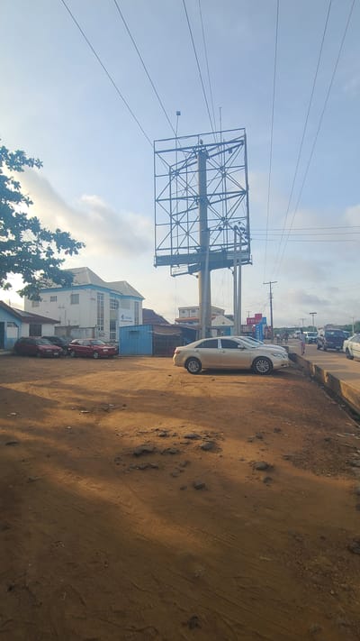 The image shows a street scene in Mandate Estate, Ilorin, Nigeria. In the foreground, there's a dirt road with tire tracks and scattered rocks. Parallel to the road, several parked cars are visible, including a light-colored sedan and multiple red sedans. Behind the cars, a building with multiple windows and a blue roof detail is present. A large, empty billboard frame stands tall, supported by a network of metal beams. Power lines stretch across the sky above the frame. The sky is partly cloudy, with visible sunshine.