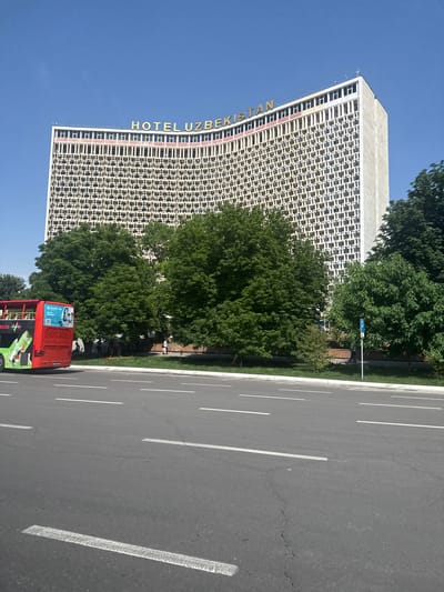 The image depicts a street scene in Kashgar mahalla, Uzbekistan, dominated by the tall, rectangular Hotel Uzbekistan building. The building's facade features a grid-like pattern and the hotel's name is vertically displayed on its side in yellow lettering. Adjacent to the hotel, lush green trees provide a contrast. A road runs alongside the hotel and trees, with clearly marked white lane dividers. A red and green tour bus is positioned on the roadside. The sky is clear and blue, suggesting a sunny day. No people are readily visible in the image.