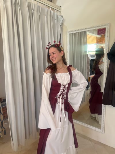The scene depicts a young adult female posing indoors, within what appears to be a changing room or costume shop in Thành phố Phan Thiết, Vietnam. The female, with long brown hair and light freckles on her cheeks, is smiling and looking towards the left of the frame. She wears a gold-colored headpiece adorned with red berry-like elements and white floral accents, along with a small, light-colored stud earring in her right ear and a thin gold chain necklace. Her attire consists of a white, off-the-shoulder blouse with long, wide, bell-shaped sleeves and a ruffled neckline. Over this, she wears a dark red (maroon) sleeveless dress or bodice featuring a white laced front closure with brown ties, resembling a corset. A tiered white skirt is visible beneath the red overlay. To the left, light grey pleated curtains hang from a track. To the right, a large white ornate framed mirror reflects the subject's back, an outdoor brick wall, a white air cooler unit, and a dark garment hanging on the far right. The floor is light-colored, and a patterned textile is partially visible on the far left.