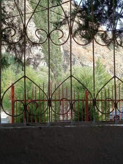 The image shows a view through a decorative metal fence, with a mountain landscape and vegetation in the background. The fence is made of dark metal with ornate scrollwork and vertical bars. In front of the fence, there is a concrete structure at the bottom of the frame. Behind the fence, there are lush green trees and bushes, and a rocky hillside rises in the distance. The sky is visible at the top. The scene appears to be outdoors, possibly in a residential area or a park, given the presence of the fence and cultivated greenery. The specific location in the city of Varzob, Tajikistan, is suggested by the context, but the image itself does not contain any explicit textual or symbolic identifiers of that location. There are no people or animals visible in the frame. The lighting suggests it is daytime, likely with bright sunlight given the highlights and shadows.