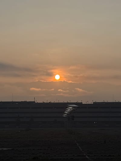 This is a vertical, medium-shot photograph of a sunrise over a city landscape. The sun, a bright orange orb, is partially obscured by scattered clouds in the upper center of the frame. The sky is a gradient of pale orange and beige. Below the sky is a dark, multi-story building, likely a parking garage, with multiple levels and rows of lights along its top. In front of the building, a field of dark, dry vegetation stretches across the foreground. A series of white, angled solar panels are visible on the left side of the frame, casting shadows on the ground. The scene appears to be captured from a distance, with the city structures forming a silhouette against the dawn sky. The location is identified as San Miguel Totoltepec, Mexico.