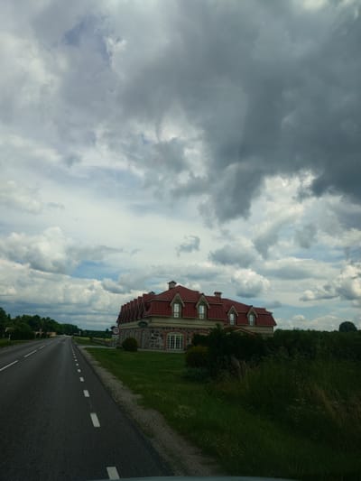 A road in the city of Pikva, Estonia, leads past a large building with a red roof. The road is paved and marked with white lines. The building is made of stone and has many windows. The sky is cloudy. There are trees and grass in the background.