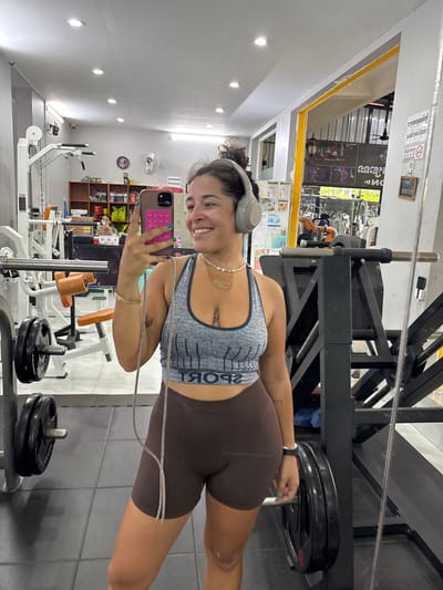 A young woman with dark curly hair pulled up, wearing a grey sports bra and brown athletic shorts, takes a mirror selfie in a gym located in Kampot, Cambodia. She is wearing large grey headphones and a necklace. She holds a pink phone with a pop socket in her right hand. The gym appears to be a small, local facility with weightlifting equipment, including barbells and weight plates, visible in the background. The walls are painted white and grey, with some yellow accents. There are shelves with products and signage in Khmer script on the walls. The floor is tiled.