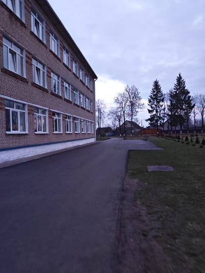 The image shows a three-story brick building with multiple windows located in Krychau, Belarus. The building is situated next to a paved walkway. Beyond the walkway is a grassy area with scattered trees and shrubs. In the distance, there are houses and a cloudy sky. The light suggests it is either early morning or late evening. There are no people visible in the scene.