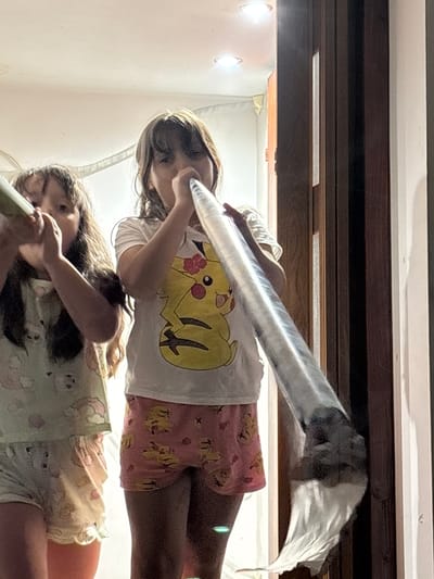 Two young girls are shown in a close-up, low-angle shot. The top girl, wearing a white t-shirt with a yellow Pikachu design and pink shorts with a similar pattern, is holding a long, clear plastic tube to her mouth, as if blowing into it. Her eyes are focused on the tube. Below her, another girl, wearing a light-colored pajama set with a floral print, is also positioned as if she is about to interact with the tube. Her face is partially visible.

The setting appears to be indoors, possibly a bedroom or play area, with soft lighting coming from overhead fixtures. The background shows wooden paneling, suggesting furniture or shelving. The overall impression is one of playful activity. The image is taken from a perspective that looks upwards at the subjects. There is no visible text in the image. The time of day and weather are not discernible. The emotion conveyed is one of childhood innocence and engagement in play. The location context is Enevo, Bulgaria.