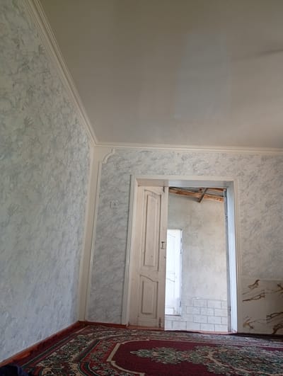 The image depicts an interior view of a room, likely in a residential building. The walls are decorated with a marbled wallpaper, featuring a pattern of grey and white swirls. A white ceiling with ornate molding is visible at the top. In the center of the image, a white wooden door is ajar, revealing another doorway beyond. This inner doorway leads to a space with a tiled floor and walls, and a partially visible second door. Exposed wooden beams suggest unfinished construction or a rustic aesthetic in the inner space. A corner of a red and patterned rug is visible on the floor in the foreground. The scene is lit by natural light entering from the open doorways. There are no people visible in the image. The location is identified as the city of Hisor, Tajikistan.