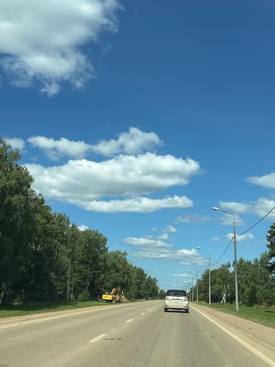 The image depicts a view from a vehicle driving on a two-lane asphalt road through a rural or suburban area, possibly the city of Ознобишино, Russia. The road is lined with trees on both sides, with lush green foliage. The sky is a clear blue with scattered white cumulus clouds. On the right side of the road, there are several streetlights and utility poles. In the distance, on the left side of the road, an excavator is visible near a pile of dirt, suggesting ongoing construction or road work. A white car is driving ahead of the viewpoint vehicle. The scene is illuminated by daylight.