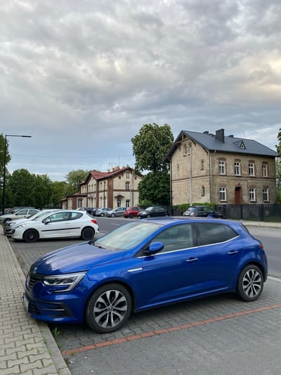 The image captures a street scene in Dobieżyn, Poland. A bright blue car is parked on a paved sidewalk, near a curb and some grass. Several other cars, including a white hatchback, are parked behind it along the street. Buildings line the opposite side of the street, including brick structures with multiple windows. Trees are scattered along the street and in front of the buildings. The sky is overcast with cloudy conditions. No people are visible in the image. The overall setting is an urban residential area.
