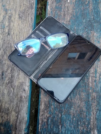 A pair of blue-rimmed eyeglasses and a smartphone are placed on a weathered, blue-painted wooden surface. The eyeglasses are open, with their lenses reflecting the sky and trees. The smartphone is closed, lying face down with its screen exhibiting minor scratches. The wooden surface appears to be part of an outdoor bench or table, with visible grain and chipped paint. The background shows glimpses of foliage and hints of the urban environment of Vlasivka, Ukraine.