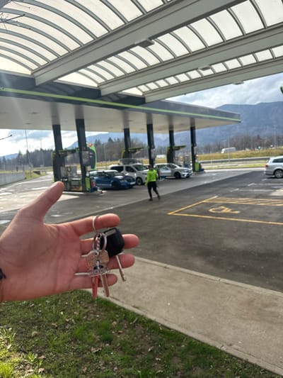 The scene depicts an outdoor fuel station located in Radovljica, Slovenia, under daylight conditions with a partly cloudy sky.

In the immediate foreground, a light-skinned left hand is visible, holding a keyring. The keyring contains a black plastic car key fob, two brass-colored metal keys, two red-colored metal keys, a silver-colored metal key, a small keychain with a portrait image, and a keychain partially displaying the text "HERO." A dark-ink tattoo is present on the inner wrist of the hand. The hand is positioned above a patch of green grass with sparse brown foliage.

The midground features a multi-lane fuel station with a translucent, ribbed canopy supported by dark pillars accented with a green stripe. Several vehicles are visible: a blue sports car, a white SUV equipped with a roof rack, a silver sedan, and other white and silver vehicles parked or in motion. Green and dark-colored fuel pumps are positioned at various bays. A person wearing a bright green long-sleeved top and dark trousers is walking from left to right across the asphalt ground, with their back to the camera. A yellow-outlined disabled parking space symbol is clearly marked on the asphalt. A concrete curb separates the grassy foreground from the asphalt area.

In the background, a road or highway is visible, with a white vehicle present. Distant trees line the horizon, beyond which large mountains rise against a grey, cloudy sky.