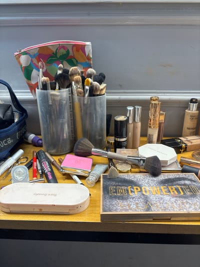 A collection of makeup products is arranged on a wooden surface, likely a vanity or table. In the background, a floral patterned makeup bag sits behind two clear cups filled with makeup brushes. Several makeup bottles and tubes, including foundation and concealer, are scattered across the surface. Also visible are various makeup brushes, an eyeshadow palette, and a compact makeup case. The scene appears to be an indoor setting, with a gray wall in the background. The lighting suggests it is daytime. There are no people present in the image. The location in Sweileh, Jordan, is not discernible from the visual evidence in the image.