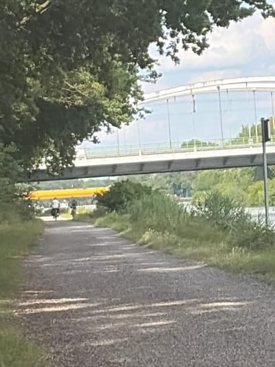 The image captures an outdoor scene with a paved path in the foreground, bordered by greenery and trees. In the mid-ground, there is a bright yellow structure, possibly a bridge or underpass, with a modern, glass-enclosed walkway above it. Two cyclists are visible on the path, heading away from the viewer, with one appearing to be slightly ahead of the other. The sky above is cloudy and light blue. The overall impression is of a sunny day, possibly late afternoon, given the shadows. The location cues suggest Garbsen, Germany, with the distinctive architecture of the overpass. The image is oriented vertically and appears to have been taken from a slightly elevated perspective. No text is visible in the image.