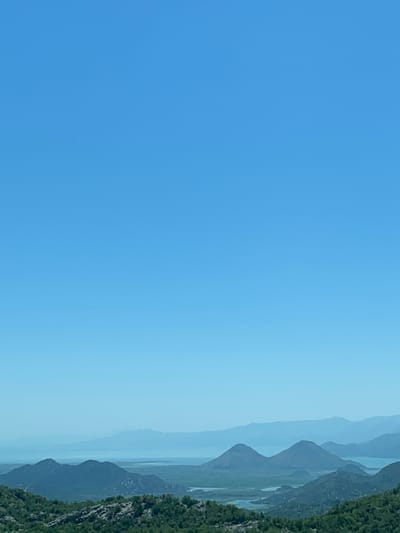 The image depicts a landscape scene with a vast expanse of a clear, bright blue sky dominating the left side. On the right side, a series of hazy, layered mountains recede into the distance, rendered in muted shades of blue and gray, suggesting atmospheric perspective. The foremost mountain range on the right is covered in dense, dark green vegetation, appearing rugged and uneven. The overall impression is one of a serene, natural environment under a clear sky, likely during daylight hours. No people, objects, text, or specific activities are visible. The scene evokes a sense of open space and the grandeur of nature. Given the location context of Gornja Brova, Montenegro, the image likely captures a view of the mountainous terrain characteristic of the region.