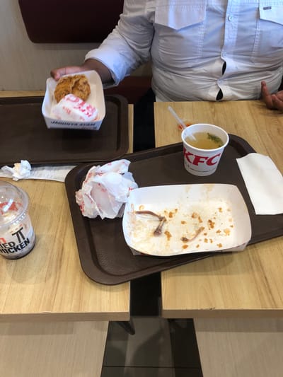 The image displays an indoor scene, likely a fast-food restaurant in Triengmatangubi, Indonesia, featuring two people at light wooden tables with KFC meals. The perspective is from above, looking down at the tables.

One person, wearing a white long-sleeved shirt, is visible from the chest down, holding a white cardboard box with a piece of fried chicken and wrapped food (likely rice) inside. The box has "KFC" branding and "JAGONYA AYAM" (Indonesian for "Champion Chicken") printed on it. This box rests on a brown tray. Another person's hand is partially visible in the upper right corner, pointing towards the table.

On a second brown tray, a meal appears to be finished. It holds a mostly empty white cardboard container with chicken bones, crumbs, and rice residue. Beside it, a white KFC-branded paper cup contains a light soup with green herbs and a plastic spork. A crumpled white napkin and other crumpled paper packaging are also on this tray.

On the wooden table, beneath the right tray, lies a long white receipt, a small crumpled napkin, and an overturned clear plastic cup. The cup has "KFC FRIED CHICKEN" and additional red text visible on its side. The overall scene depicts the aftermath or ongoing consumption of a fast-food meal, with packaging and remnants scattered on the trays and table. The tables are made of light wood, and the flooring is a mix of light and dark tiles.
