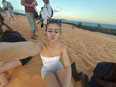 The image displays a group of individuals on the Mũi Né Red Sand Dunes in Thành phố Phan Thiết, Vietnam, at dusk. In the foreground, a young female subject with light skin and dark hair is capturing a selfie. She wears a white strapless top, light grey shorts, and sunglasses on her head, with her eyes closed and lips pursed. Her right arm is extended to hold the camera.
To her immediate left, another individual wearing a black garment is partially visible. Behind them, multiple people are present: one individual in a red t-shirt, khaki capri pants, and dark sandals holds a silver can and wears a black shoulder bag. Another person wears a white shirt and light pants, carrying a blue bag. A straw hat rests on the sand near this group. To the right of the main subject, a person with dark skin and braided hair, wearing a dark blue top, is partially visible, with a black bag or shoes on the sand.
The scene is dominated by undulating reddish-orange sand dunes, exhibiting wind-formed ripples, which slope down towards the horizon. In the mid-ground, several small figures are discernible on the distant parts of the dunes. The background reveals a calm, dark blue ocean, separated from the dunes by a strip of green vegetation and buildings with red-tiled roofs. The sky is clear, transitioning from light blue overhead to a faint yellow-orange hue near the horizon.